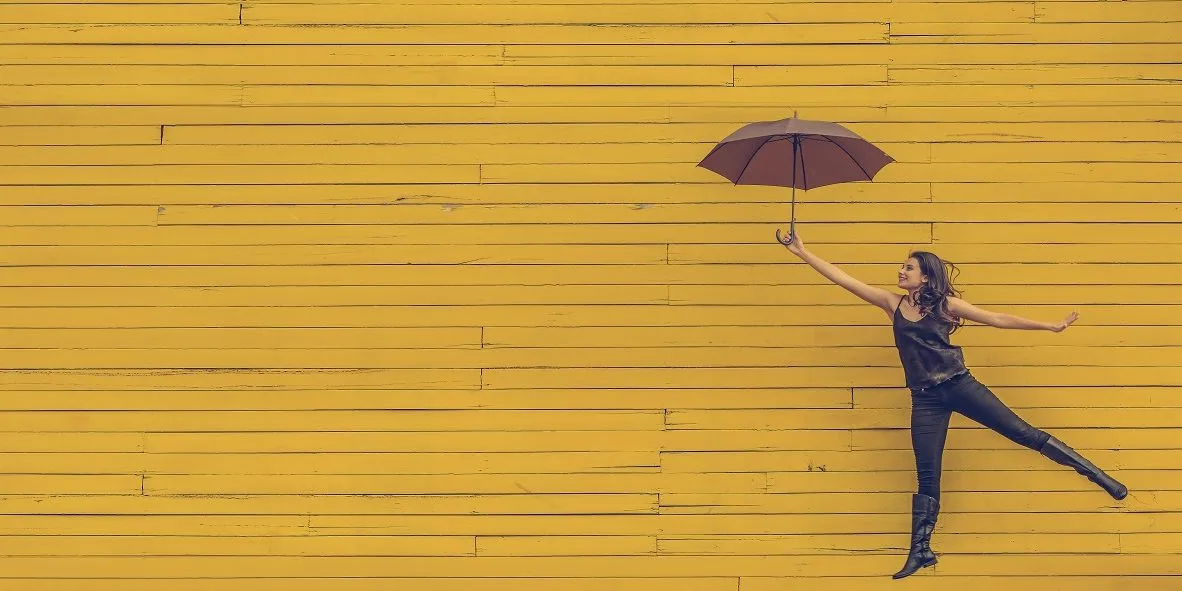 edu-lauton-TyQ-0lPp6e4-unsplash woman in black with umbrella flying across a yellow wall symbolising benefits of executive coaching